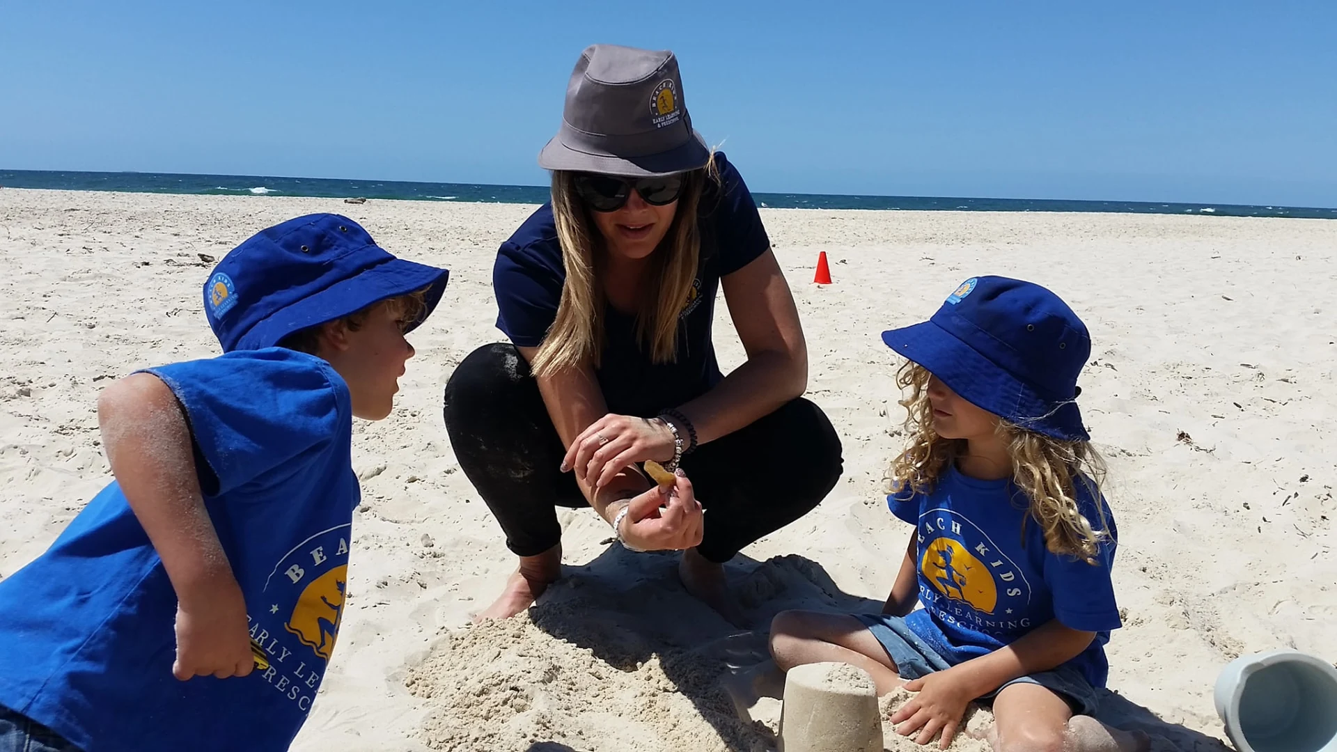 A Beach Kids educator showing two children a shell beside their sandcastle during Beach Kinder